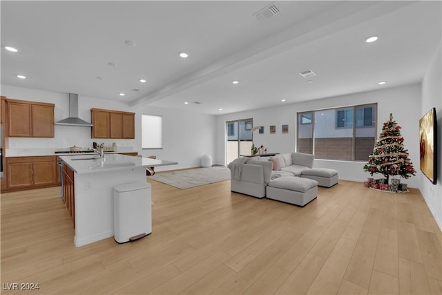 kitchen featuring wall chimney range hood, gas stovetop, beamed ceiling, a kitchen island with sink, and light wood-type flooring