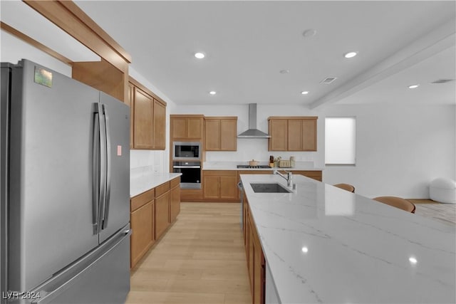 kitchen with light stone countertops, stainless steel appliances, sink, wall chimney range hood, and light hardwood / wood-style floors