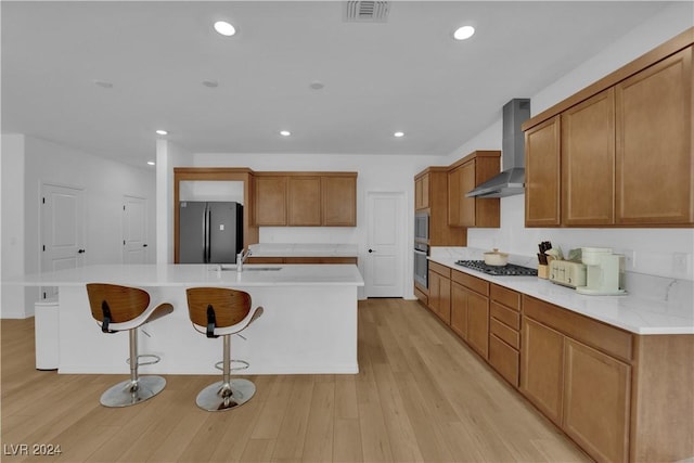 kitchen with light wood-type flooring, stainless steel appliances, a kitchen island with sink, and wall chimney exhaust hood