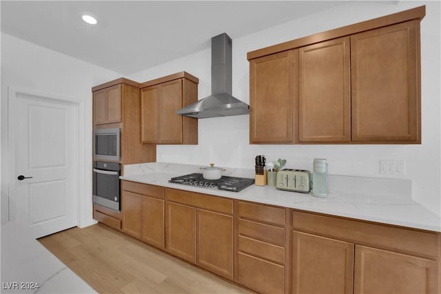 kitchen with wall chimney exhaust hood, light hardwood / wood-style floors, light stone counters, and appliances with stainless steel finishes