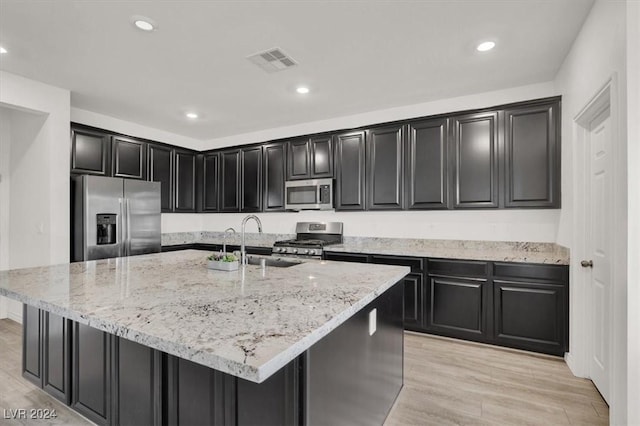 kitchen featuring sink, stainless steel appliances, light stone counters, light hardwood / wood-style flooring, and an island with sink