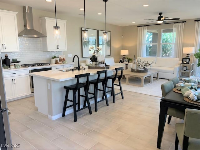 kitchen featuring tasteful backsplash, wall chimney exhaust hood, a kitchen island with sink, pendant lighting, and white cabinetry