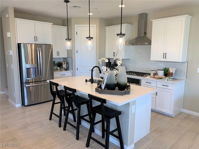 kitchen with wall chimney exhaust hood, stainless steel appliances, a kitchen island with sink, decorative light fixtures, and white cabinetry