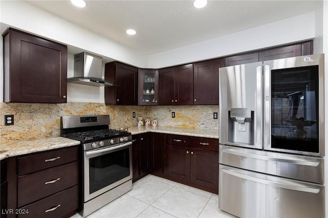 kitchen with dark brown cabinetry, light stone countertops, stainless steel appliances, wall chimney range hood, and tasteful backsplash