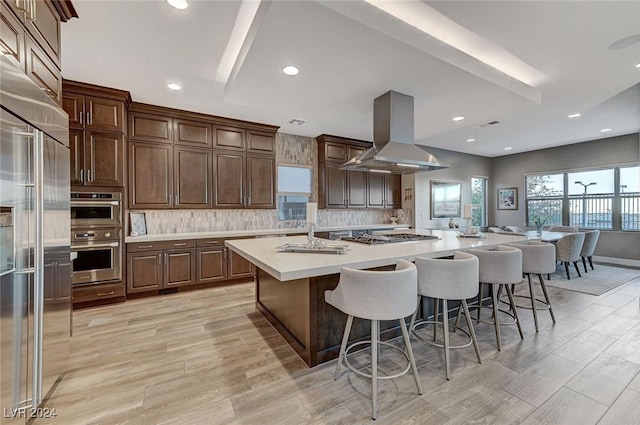 kitchen with a breakfast bar area, an island with sink, tasteful backsplash, island range hood, and stainless steel appliances