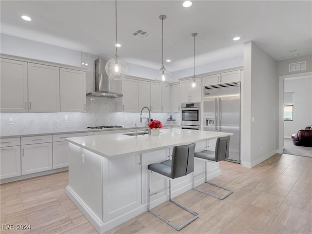 kitchen featuring appliances with stainless steel finishes, pendant lighting, sink, a center island with sink, and wall chimney exhaust hood