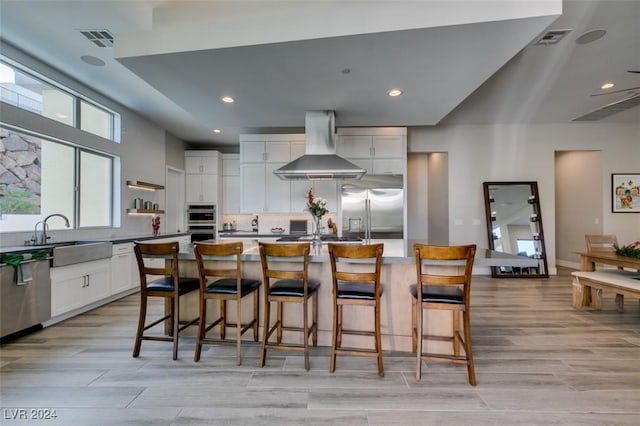 kitchen with wall chimney exhaust hood, stainless steel appliances, a kitchen island with sink, white cabinets, and a breakfast bar area