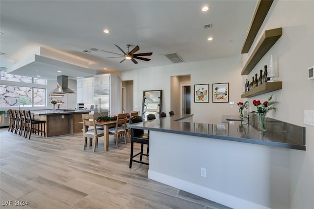 kitchen featuring kitchen peninsula, light wood-type flooring, wall chimney range hood, white cabinets, and a breakfast bar area