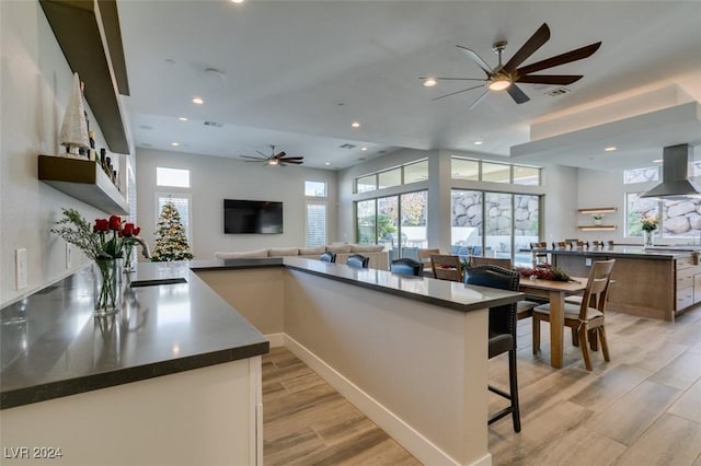 kitchen featuring a kitchen breakfast bar, wall chimney range hood, sink, ceiling fan, and light hardwood / wood-style floors