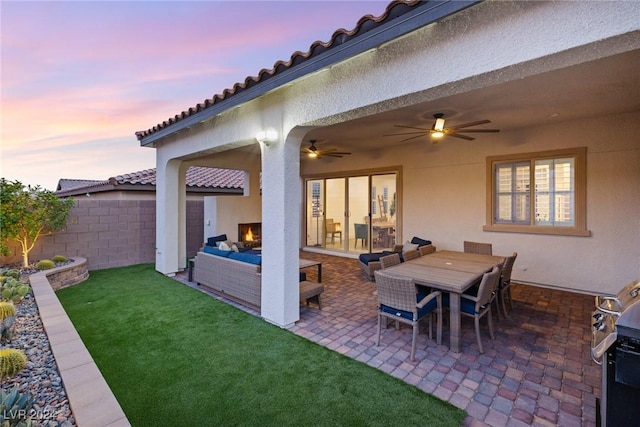 patio terrace at dusk featuring an outdoor living space with a fireplace, ceiling fan, and a yard