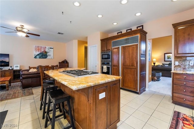 kitchen featuring light tile patterned flooring, built in appliances, a center island, ceiling fan, and light stone countertops