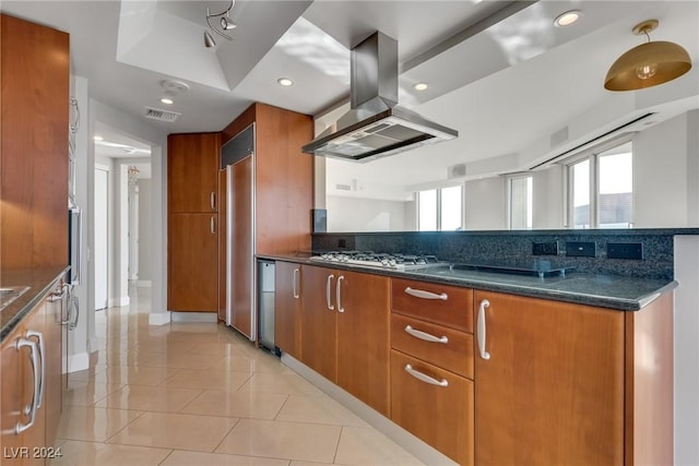 kitchen with island exhaust hood, dark stone counters, stainless steel gas cooktop, paneled built in refrigerator, and light tile patterned floors