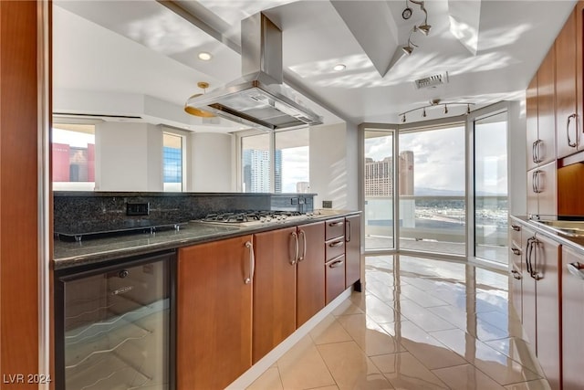 kitchen with beverage cooler, stainless steel gas cooktop, dark stone countertops, island range hood, and light tile patterned floors