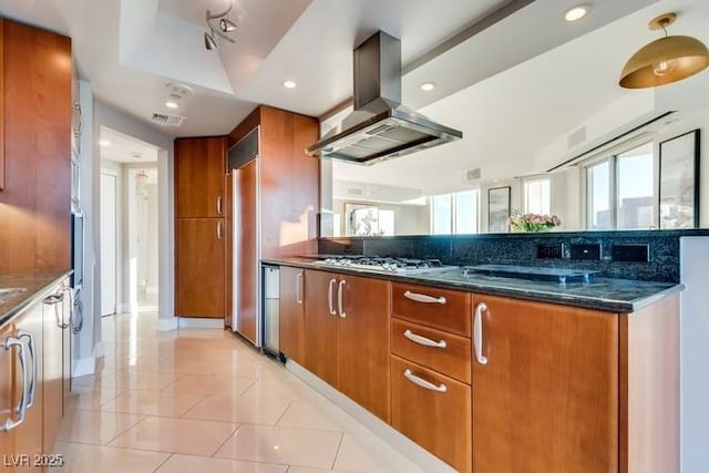 kitchen with brown cabinets, island exhaust hood, light tile patterned floors, stainless steel gas stovetop, and dark stone countertops