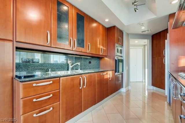 kitchen featuring brown cabinets, stainless steel microwave, backsplash, glass insert cabinets, and a sink