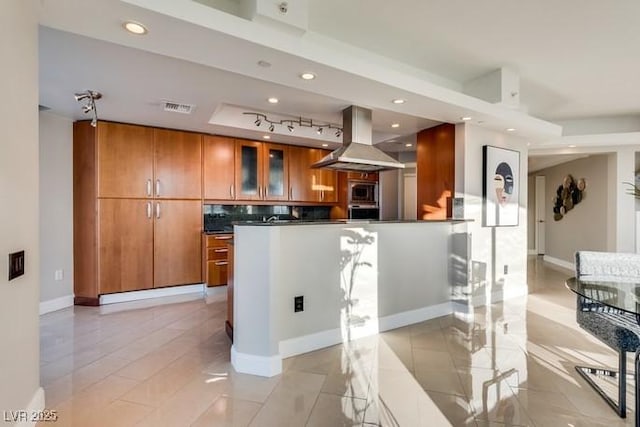 kitchen featuring visible vents, dark countertops, glass insert cabinets, brown cabinets, and island exhaust hood