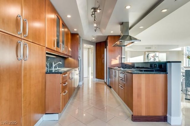 kitchen with island exhaust hood, tasteful backsplash, brown cabinetry, glass insert cabinets, and a sink