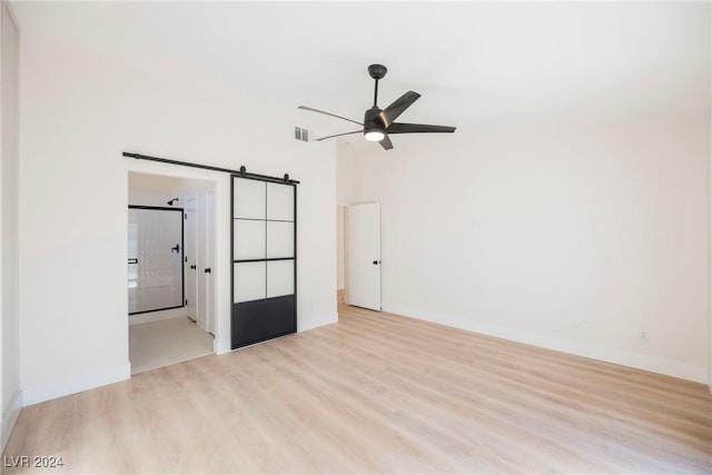 unfurnished bedroom featuring connected bathroom, ceiling fan, light wood-type flooring, and a barn door