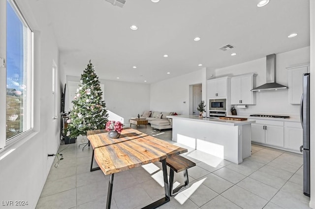 kitchen featuring wall chimney exhaust hood, light tile patterned floors, a kitchen island, white cabinets, and appliances with stainless steel finishes