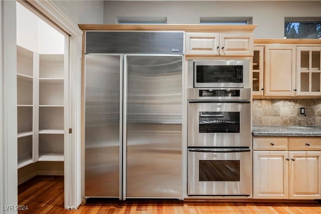 kitchen featuring built in appliances, decorative backsplash, light hardwood / wood-style flooring, and dark stone countertops