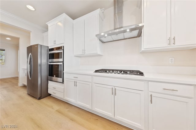 kitchen with appliances with stainless steel finishes, ornamental molding, wall chimney range hood, white cabinets, and light hardwood / wood-style floors