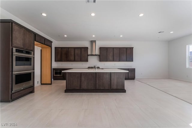 kitchen with dark brown cabinetry, a kitchen island with sink, stainless steel double oven, and wall chimney range hood