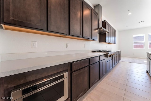 kitchen with light tile patterned flooring, dark brown cabinetry, stainless steel gas cooktop, and wall chimney range hood