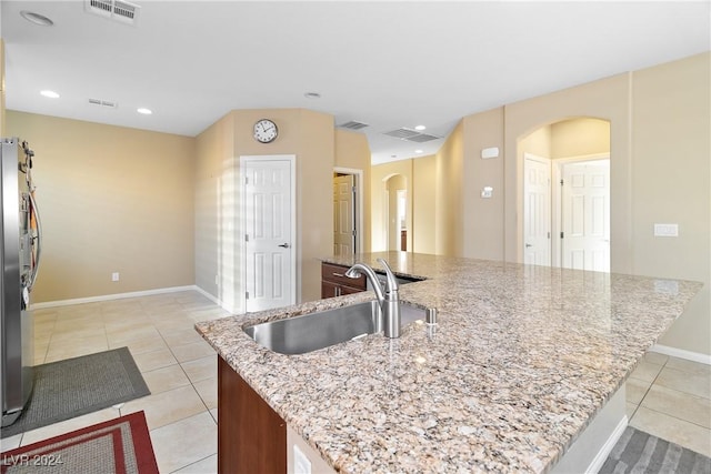 kitchen with a center island with sink, sink, stainless steel fridge, light tile patterned flooring, and light stone counters