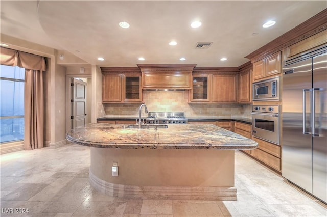 kitchen featuring sink, built in appliances, custom range hood, dark stone countertops, and a center island with sink