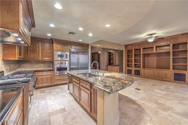 kitchen featuring built in appliances, a kitchen island with sink, dark stone counters, ceiling fan, and sink