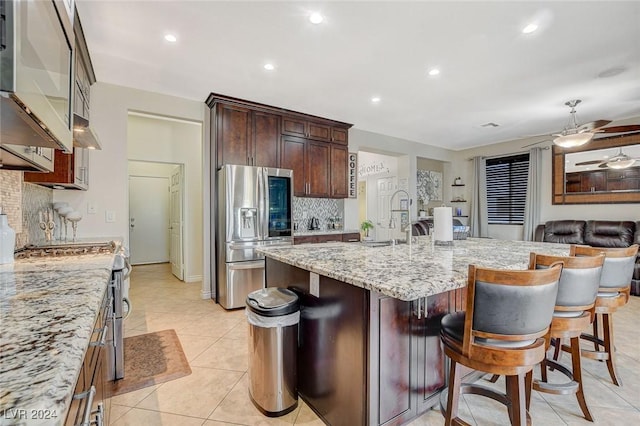 kitchen featuring backsplash, light stone counters, stainless steel appliances, a center island with sink, and a breakfast bar area