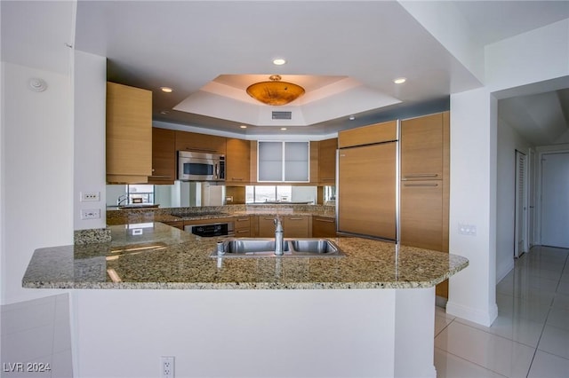 kitchen with sink, paneled built in refrigerator, a tray ceiling, light stone counters, and kitchen peninsula