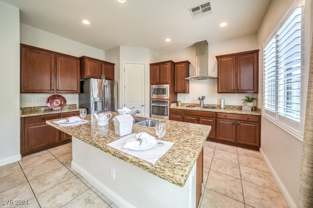 kitchen featuring light stone countertops, wall chimney range hood, a kitchen island with sink, light tile patterned flooring, and appliances with stainless steel finishes