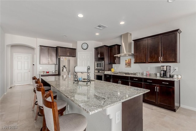 kitchen featuring light stone countertops, wall chimney range hood, a kitchen bar, a center island with sink, and appliances with stainless steel finishes