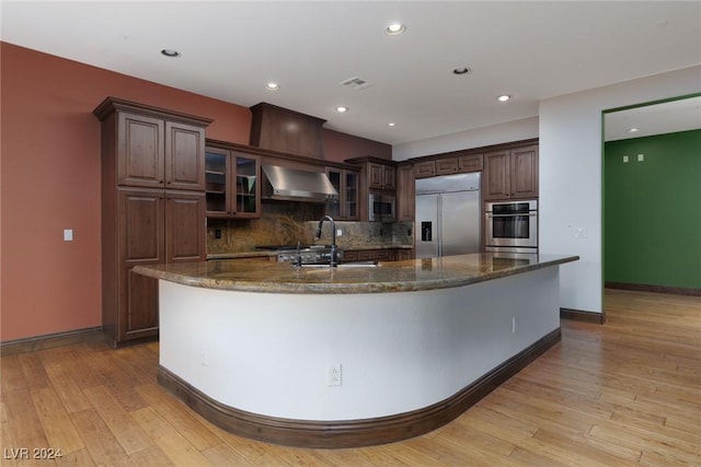 kitchen featuring backsplash, wall chimney exhaust hood, dark brown cabinets, a spacious island, and built in appliances