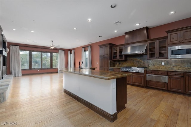 kitchen with appliances with stainless steel finishes, backsplash, ceiling fan, a kitchen island with sink, and wall chimney range hood