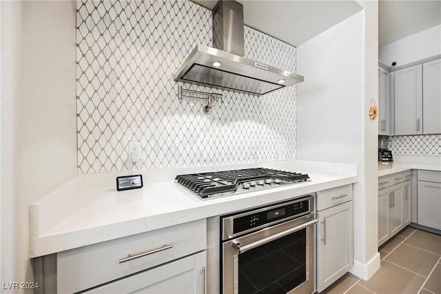 kitchen with decorative backsplash, gray cabinetry, stainless steel appliances, wall chimney range hood, and light tile patterned floors