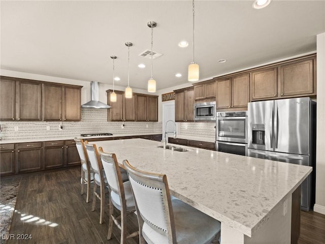 kitchen featuring wall chimney exhaust hood, pendant lighting, a kitchen island with sink, appliances with stainless steel finishes, and sink