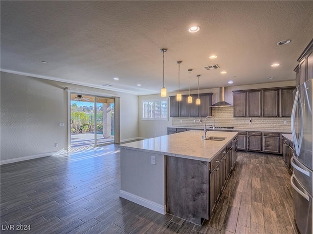 kitchen featuring stainless steel fridge, a kitchen island with sink, wall chimney exhaust hood, and pendant lighting