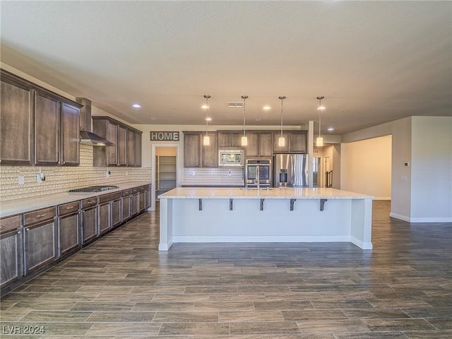 kitchen with a center island with sink, appliances with stainless steel finishes, hanging light fixtures, decorative backsplash, and wall chimney range hood