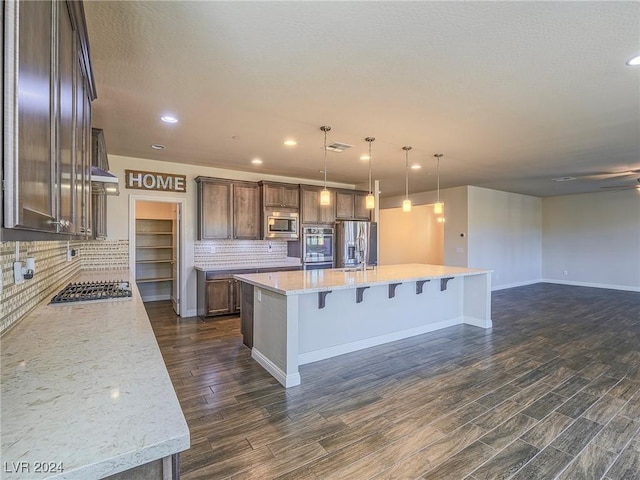 kitchen featuring stainless steel appliances, hanging light fixtures, a large island with sink, light stone counters, and backsplash