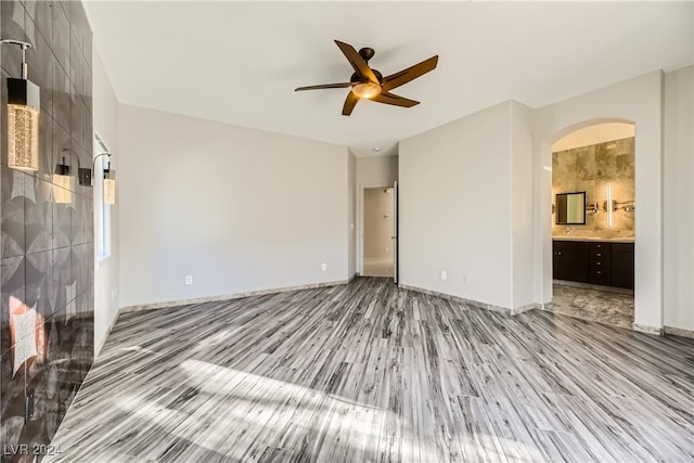 unfurnished bedroom with arched walkways, ensuite bathroom, light wood-style floors, a ceiling fan, and baseboards