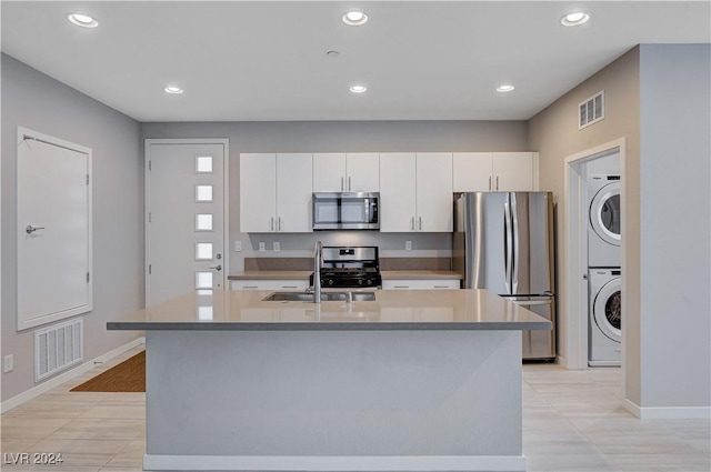 kitchen featuring a center island with sink, sink, stacked washing maching and dryer, appliances with stainless steel finishes, and white cabinets