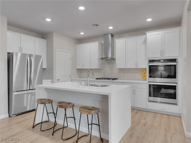 kitchen featuring white cabinetry, sink, wall chimney exhaust hood, stainless steel appliances, and an island with sink