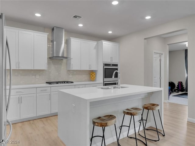 kitchen featuring light wood-type flooring, stainless steel appliances, wall chimney range hood, white cabinetry, and an island with sink