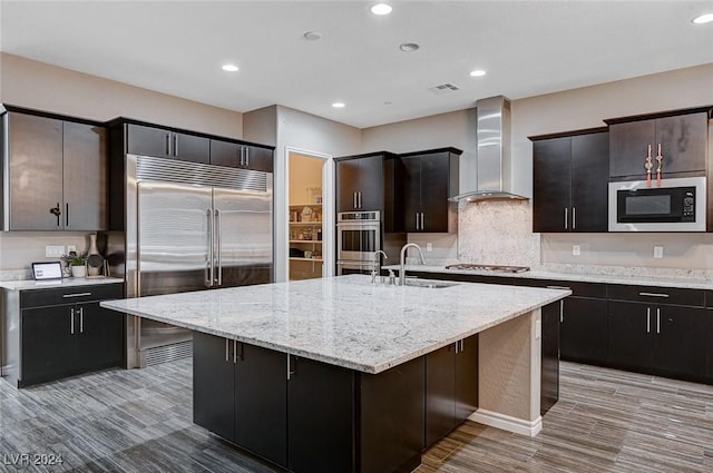 kitchen featuring built in appliances, wall chimney range hood, a kitchen island with sink, and a breakfast bar area