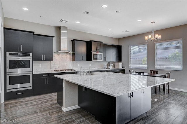 kitchen with wall chimney range hood, sink, an island with sink, appliances with stainless steel finishes, and decorative light fixtures