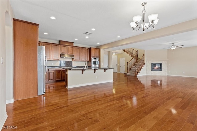 kitchen featuring wood-type flooring, hanging light fixtures, a center island with sink, stainless steel appliances, and ceiling fan with notable chandelier