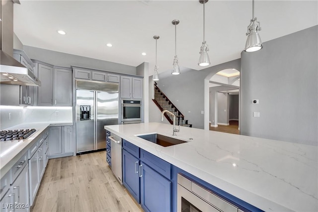 kitchen with wall chimney range hood, hanging light fixtures, sink, appliances with stainless steel finishes, and light stone counters