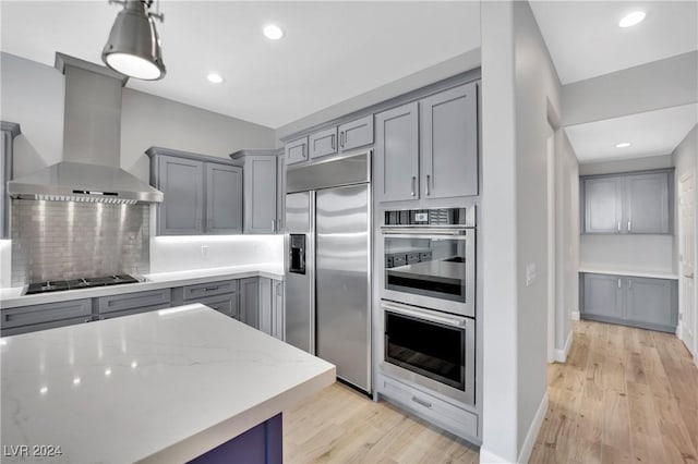 kitchen featuring appliances with stainless steel finishes, backsplash, light stone counters, gray cabinetry, and wall chimney range hood